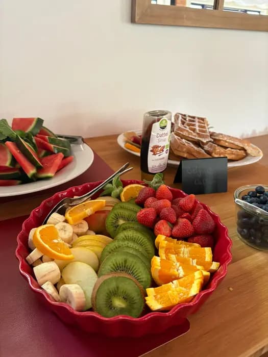 Obstschale mit Kiwi, Orange, Erdbeeren und Banane, dahinter Waffeln
